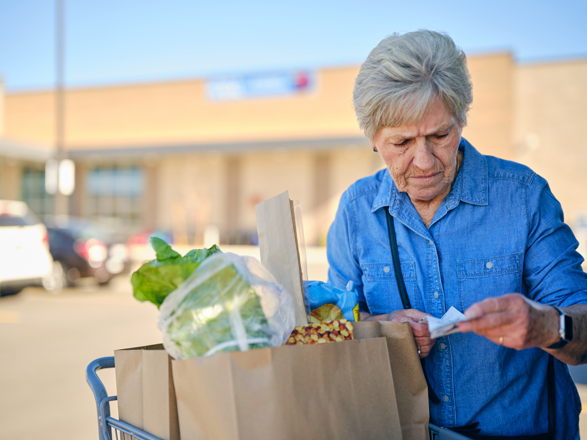 An older woman frowning at her receipt beside bags of groceries in a store parking lot