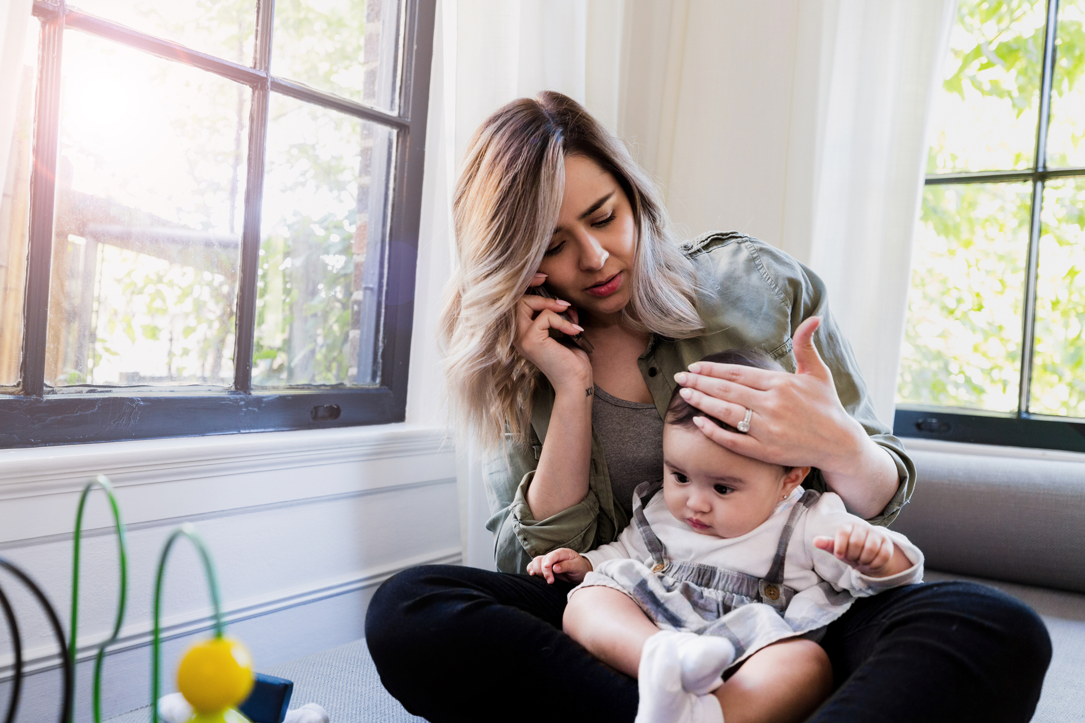 A worried mother on a phone call, resting her hand on her baby&rsquo;s head